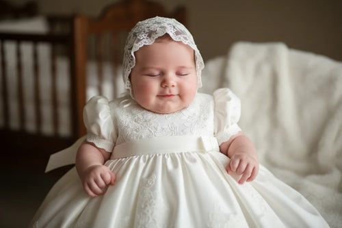 Sweet baby girl in an elegant white baptism dress, showcasing delicate lace details and a matching bonnet, representing our Elegant Baptism Dresses for Girls Collection. Perfect for special occasions and cherished memories.