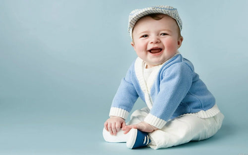 Adorable baby boy in dapper boys clothing, featuring a light blue cardigan and white pants, smiling cheerfully while sitting on a light blue background. Perfect look for a stylish young gentleman.