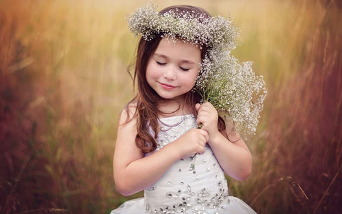 A young girl wearing a beautiful dress from the Tutus, Skirts & Dresses for Girls collection, holding delicate white flowers and wearing a flower crown, perfect for showcasing tutus and skirts in a whimsical outdoor setting.