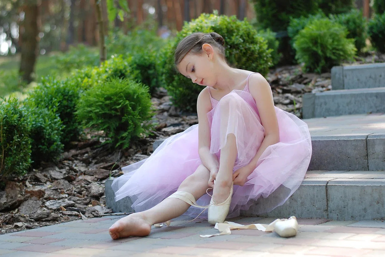 A young girl wearing ballet dance wear, sitting on a stone staircase while putting on ballet slippers, dressed in a delicate pink tutu. This charming scene captures the elegance of ballet and the joy of dance.