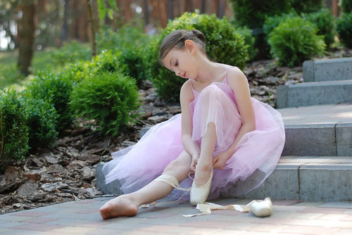 A young girl wearing ballet dance wear, sitting on a stone staircase while putting on ballet slippers, dressed in a delicate pink tutu. This charming scene captures the elegance of ballet and the joy of dance.