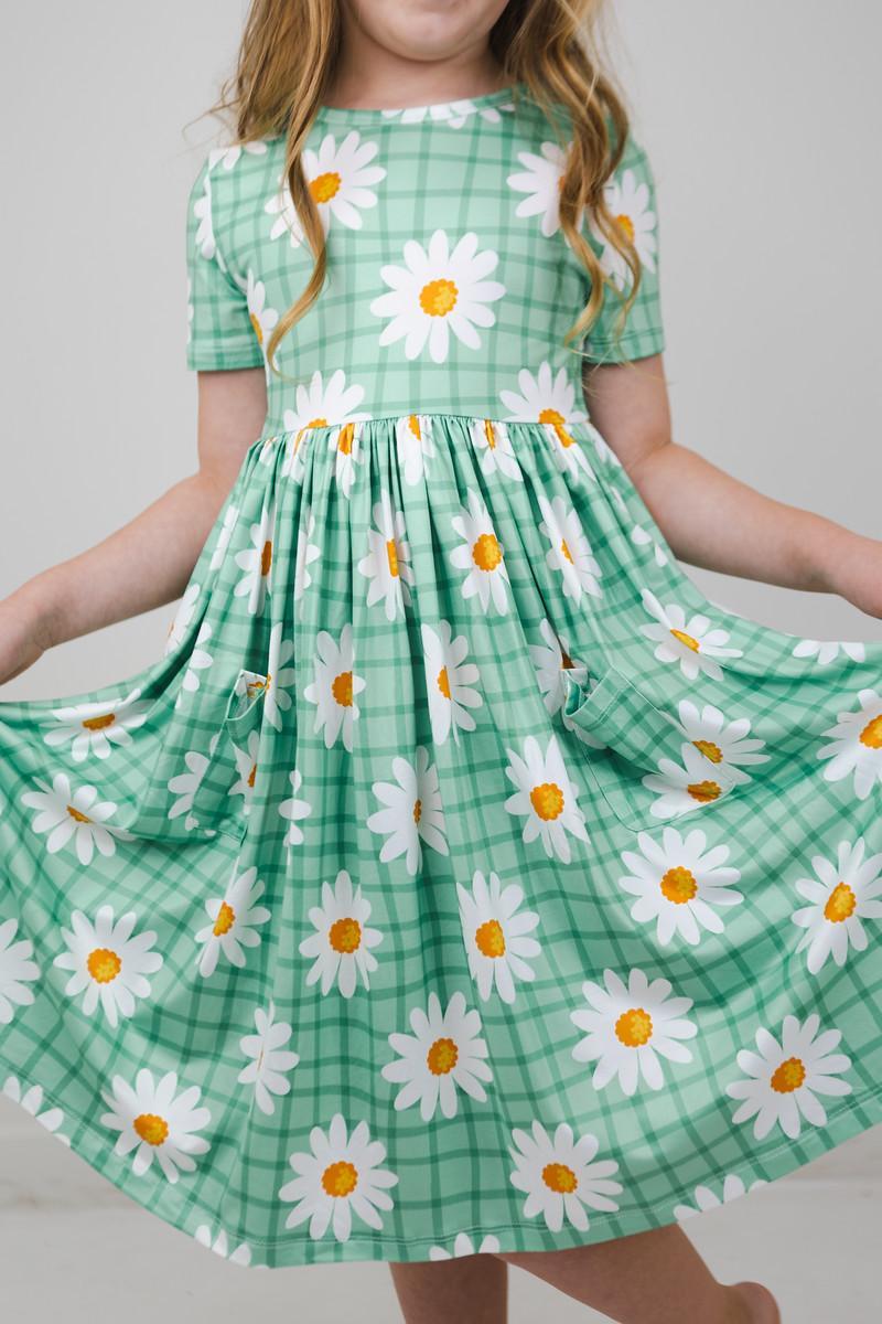 Girl twirling in the Off the Grid S/S Pocket Twirl Dress by Mila & Rose, showcasing a vibrant green design with daisies and a playful full skirt.