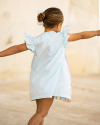 Back view of girl twirling in light blue rainbow dress with flutter sleeves, visible back zipper, and colorful pleated hem.