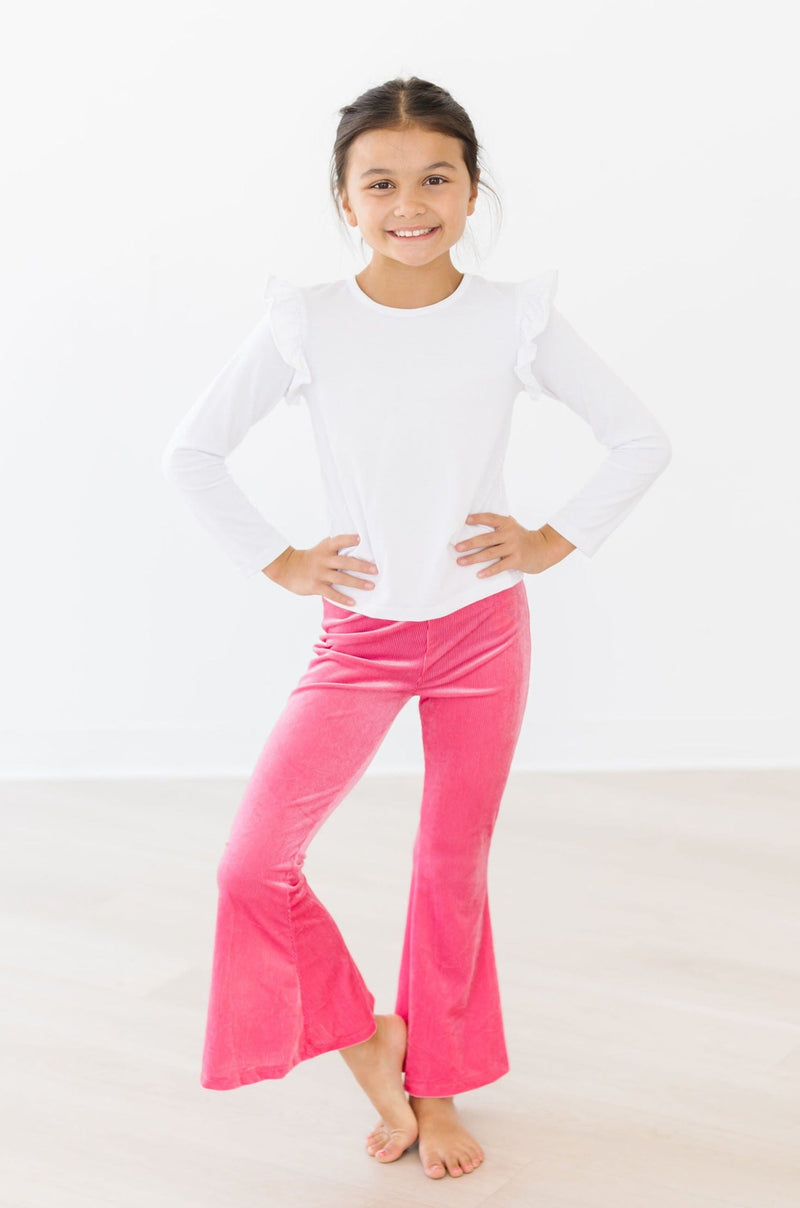 A young girl wearing Watermelon Pink Ribbed Velvet Bells paired with a white ruffled long-sleeve top, smiling and posing confidently. These stylish bell-bottoms from Mila & Rose ® feature a vibrant pink hue, perfect for fashionable kids.