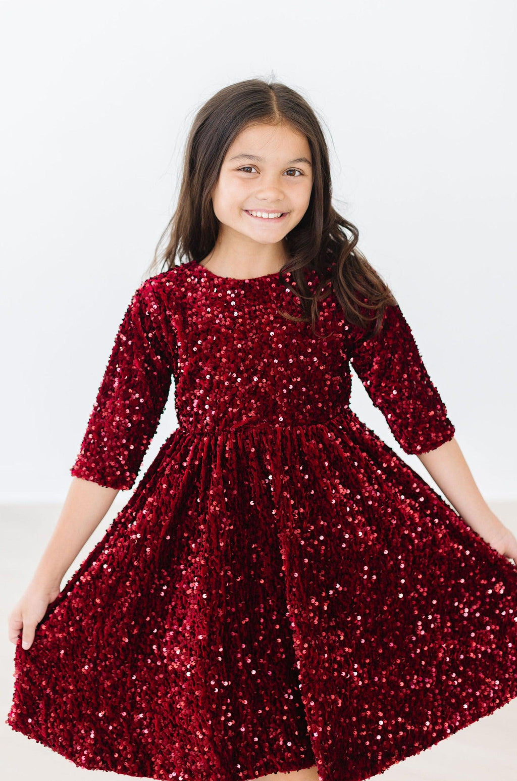 Young girl wearing a Cranberry Velvet Sequin Dress by Mila & Rose®, smiling and displaying the dress's sparkling sequins.