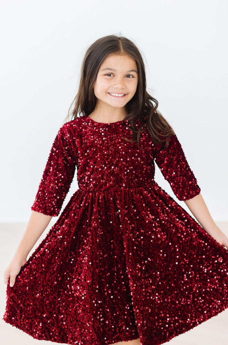 Young girl wearing a Cranberry Velvet Sequin Dress by Mila & Rose®, smiling and displaying the dress's sparkling sequins.