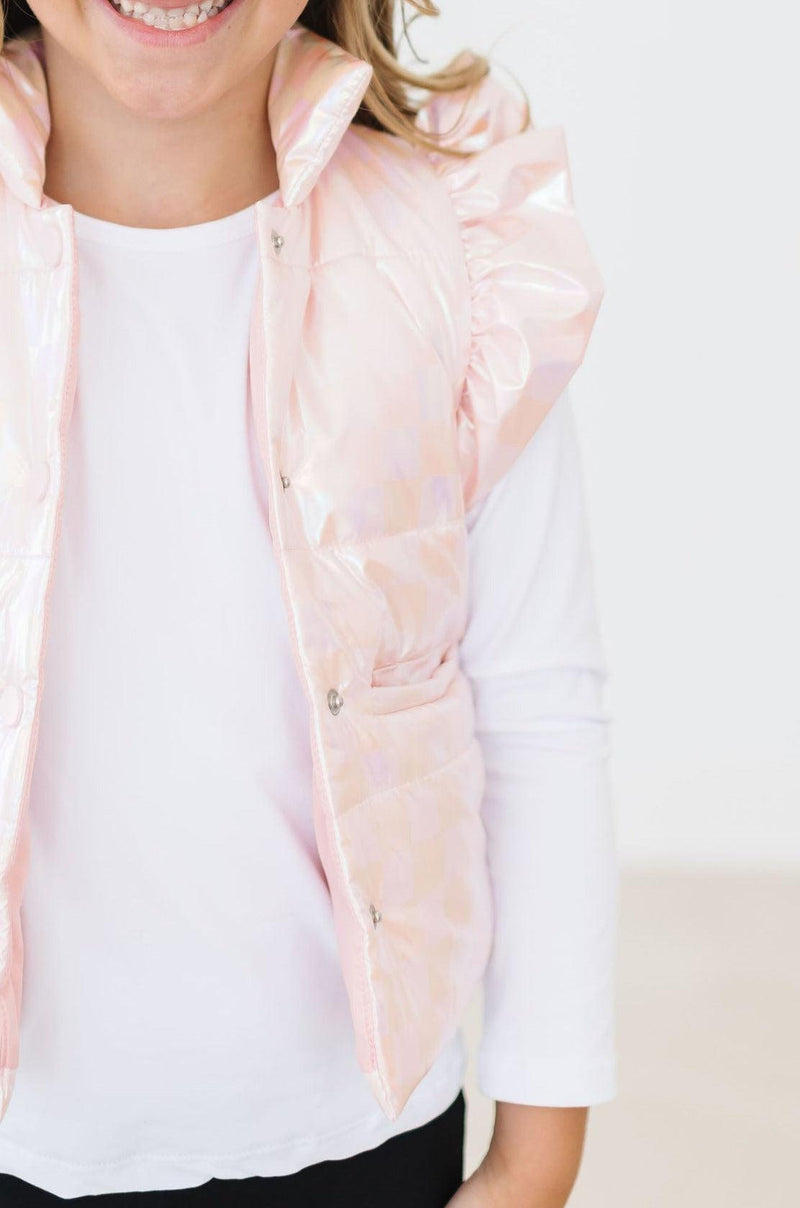 Close-up of a young girl proudly showing off her Light Pink Checker Puffer Vest with ruffled shoulders, a perfect winter layering choice.