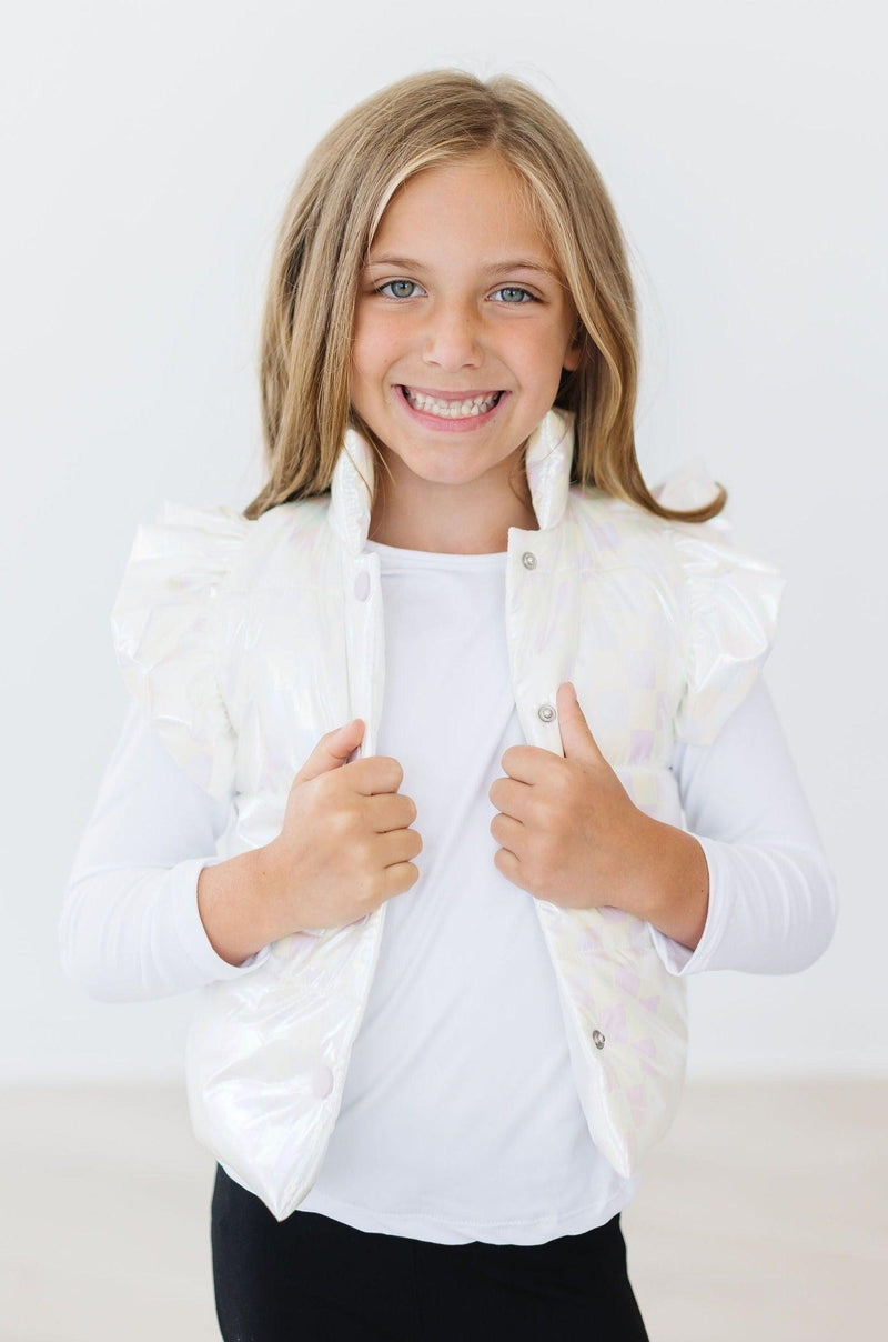 A young girl posing happily in the White Checker Puffer Vest, featuring ruffled shoulders, paired with a white long-sleeve shirt.