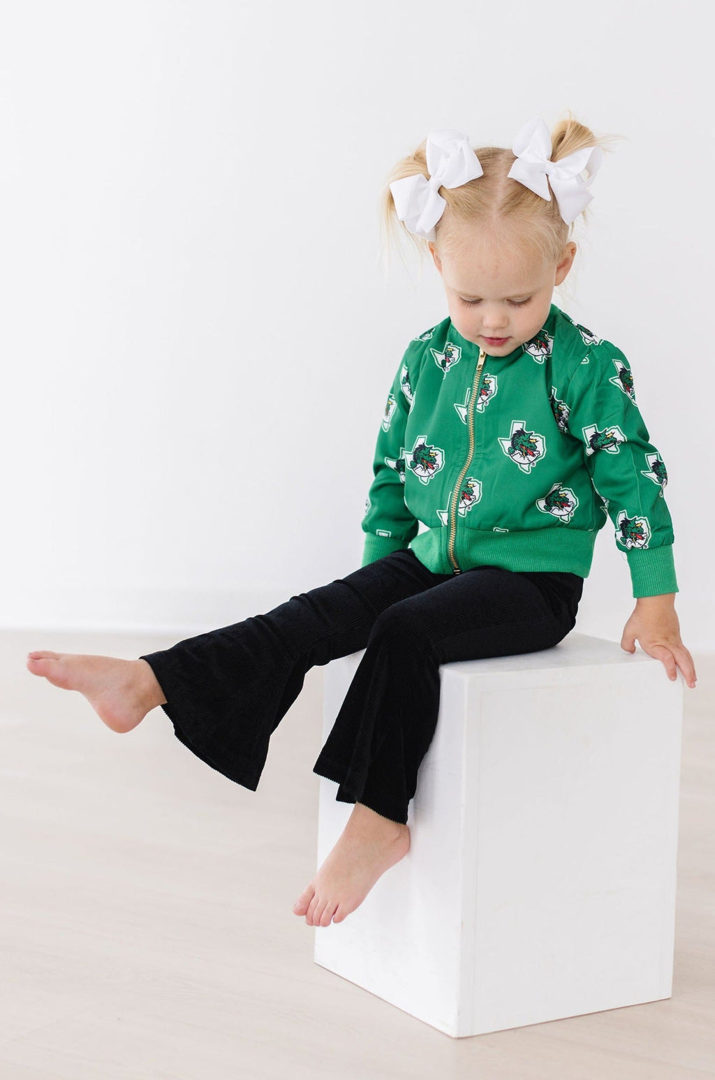 A stylish young girl wearing chic black velvet bell bottoms, paired with a green zip-up jacket and seated on a white cube, showcasing the fashionable black velvet bell bottoms for girls.