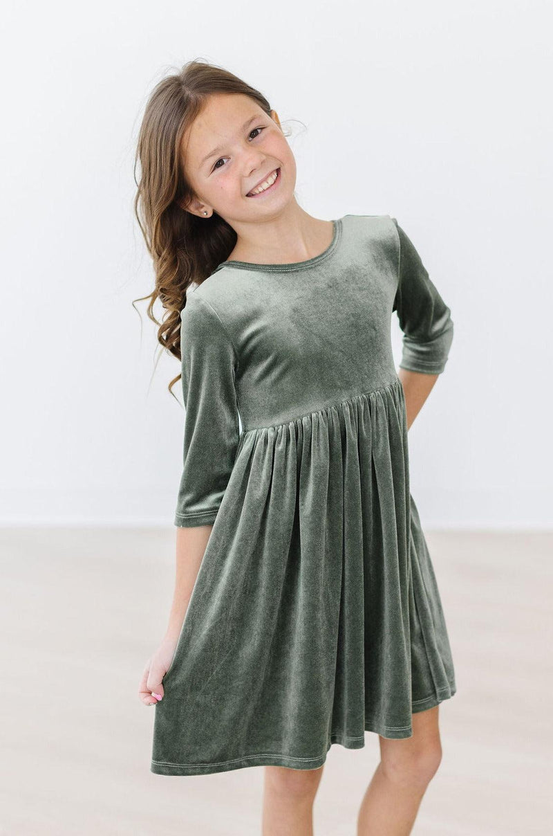 Sage Velvet Twirl Dress by Mila & Rose, showcasing a happy young girl posing in a cozy sage green dress with three-quarter sleeves, perfect for twirling and play.