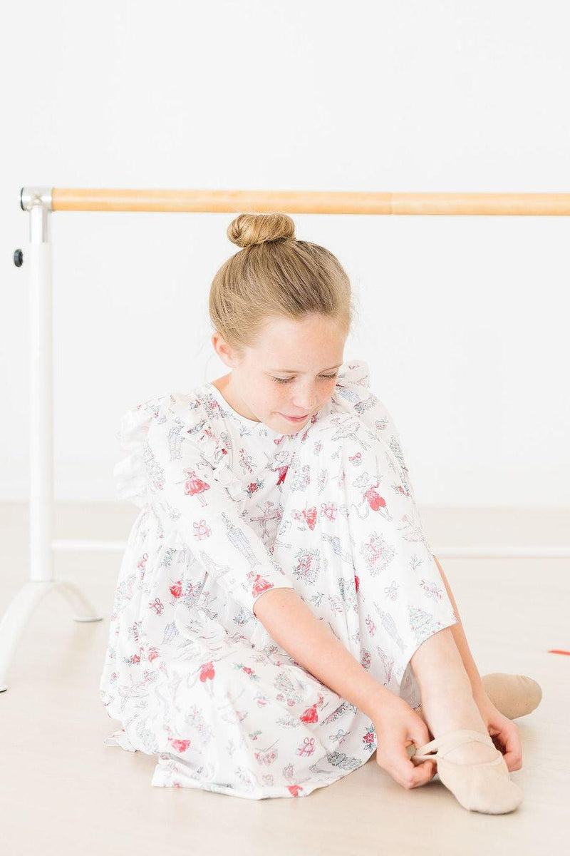 A girl wearing Clara's Christmas 3/4 Sleeve Ruffle Twirl Dress from Mila & Rose® sits on the floor, tying her ballet slippers with a serene expression.