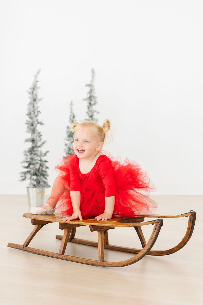 A happy toddler with pigtails sits on a rocking chair, beaming while in a Festive Red 3/4 Holiday Fluffy Tutu Leotard for Girls, capturing the joyful essence of playtime in this delightful tutu.