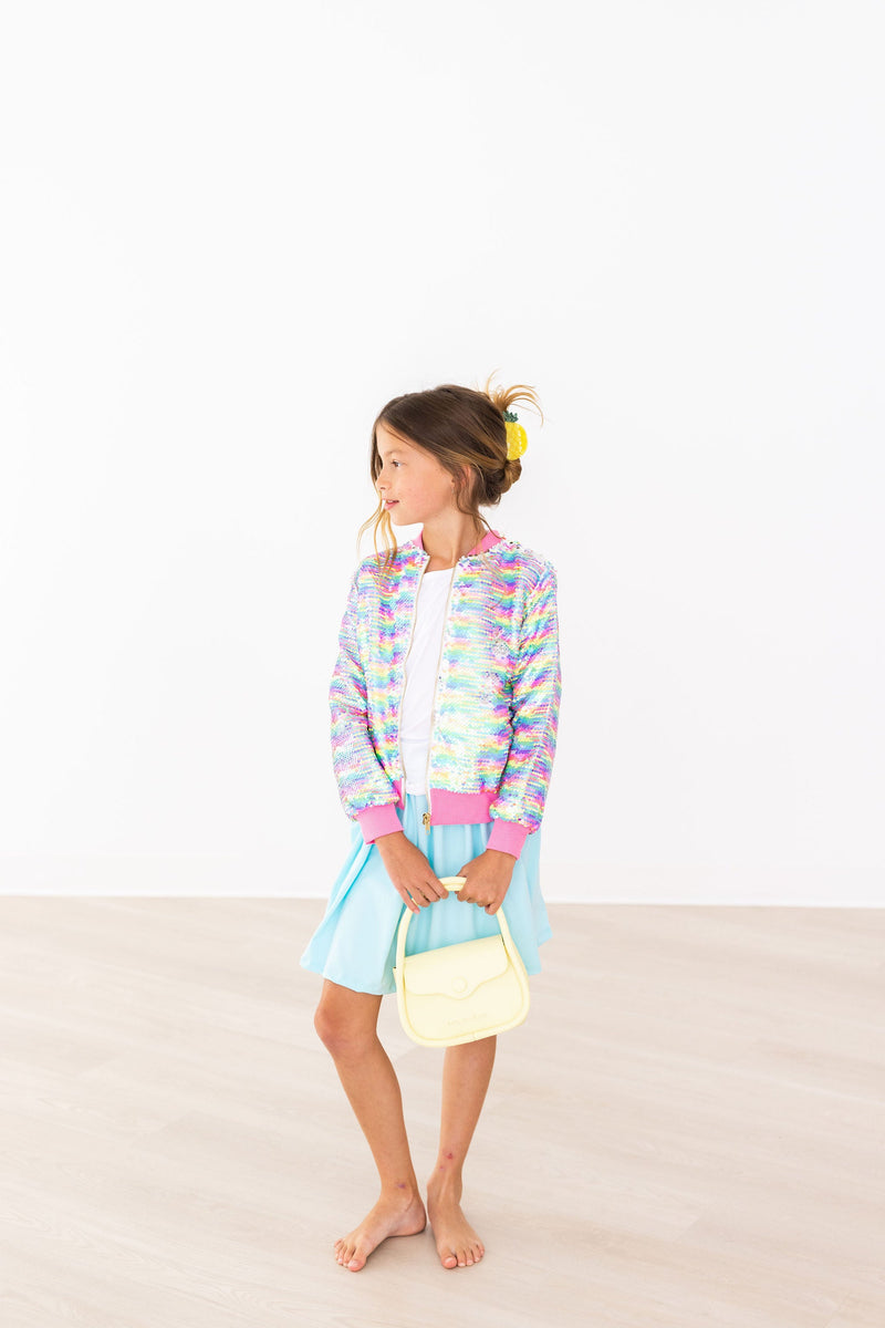 A chic young girl wearing the Aqua Twirl Skort by Mila & Rose®, paired with a colorful bomber jacket, standing confidently with a cute handbag.