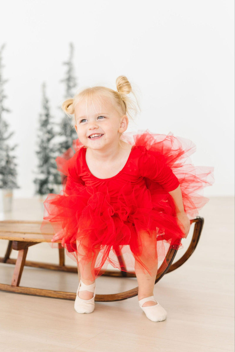 A laughing young girl dresses in a Festive Red 3/4 Holiday Fluffy Tutu Leotard for Girls, enthusiastically playing while perched on a wooden rocking chair, showcasing the fun and festive charm of the tutu.