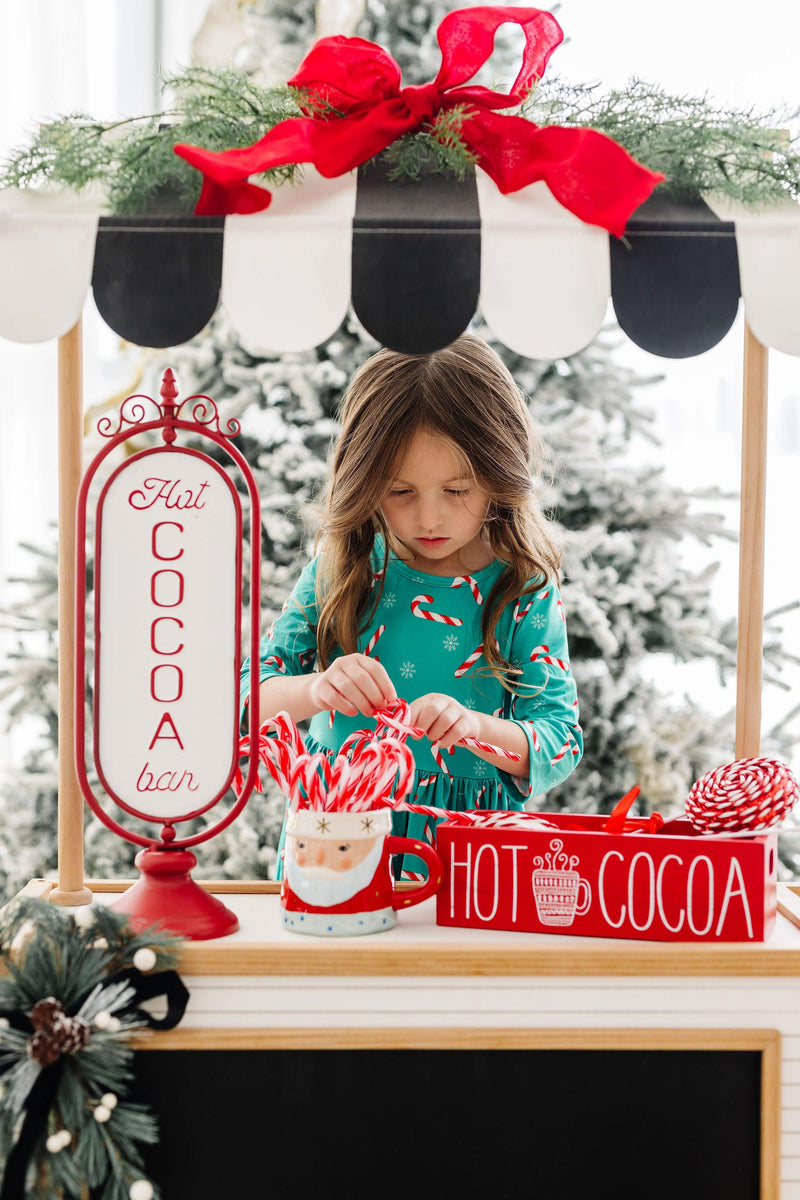 A young girl in a Christmas Candy 3/4 Sleeve Pocket Twirl Dress by Mila & Rose joyfully arranging candy canes at a festive hot cocoa bar.