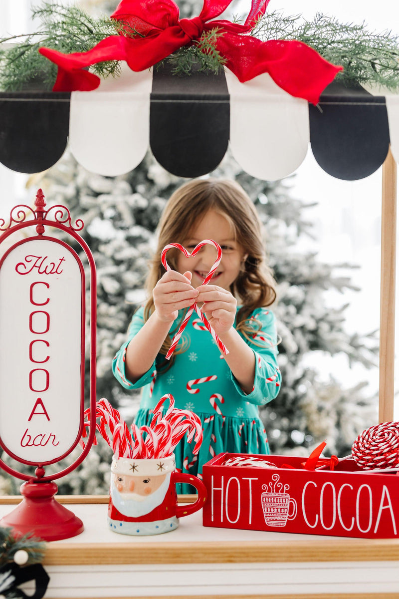 A young girl in a Christmas Candy 3/4 Sleeve Pocket Twirl Dress by Mila & Rose, holding heart-shaped candy canes at a festive hot cocoa bar.