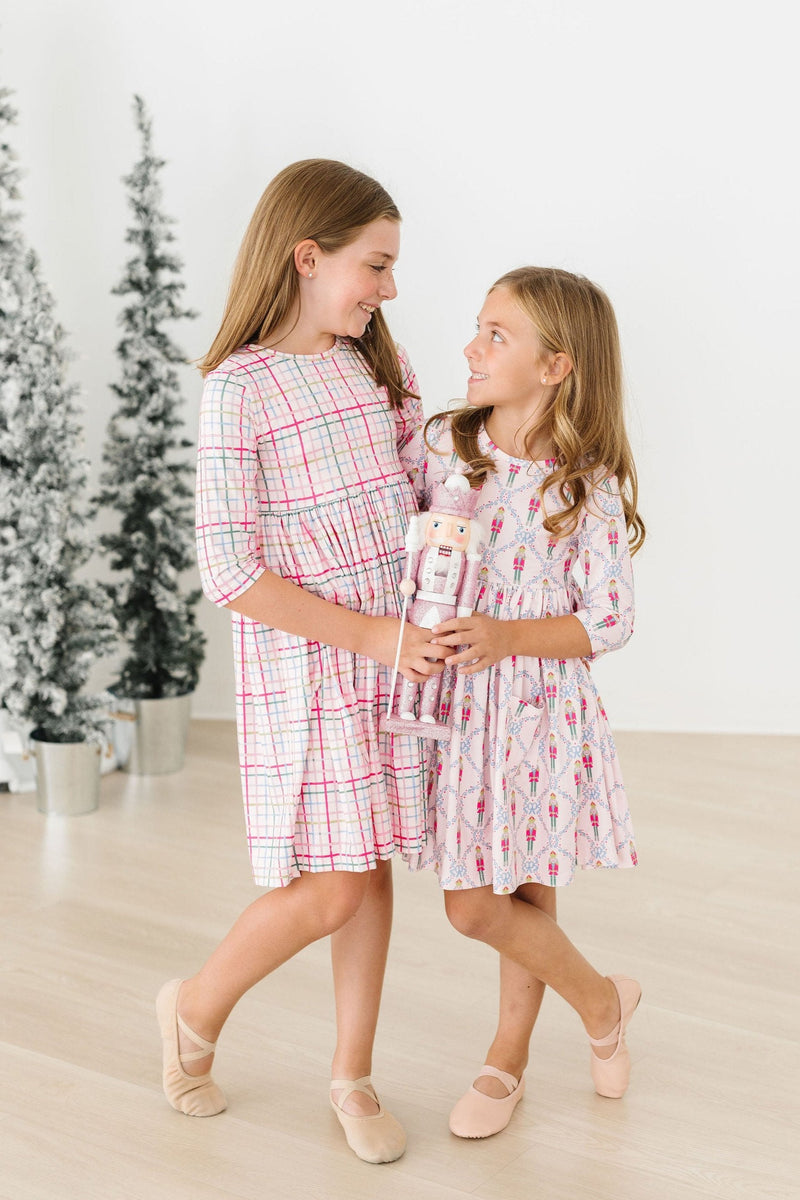 Two girls in festive attire, wearing a Pink Nutcracker 3/4 Sleeve Pocket Twirl Dress and a matching outfit, enjoying a moment together with nutcracker dolls.