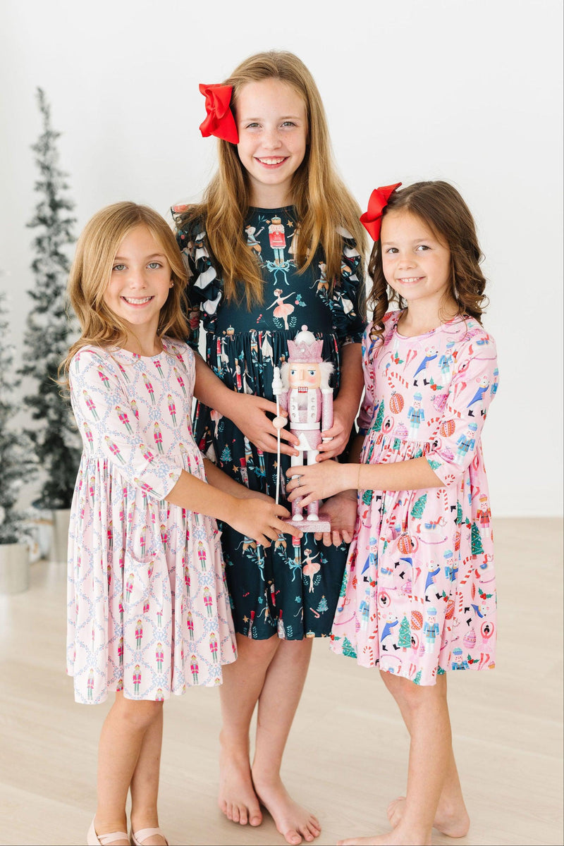 Three girls wearing the Sugarplum Fairy 3/4 Sleeve Pocket Twirl Dress in different festive patterns, joyfully holding a nutcracker against a backdrop of holiday decorations.