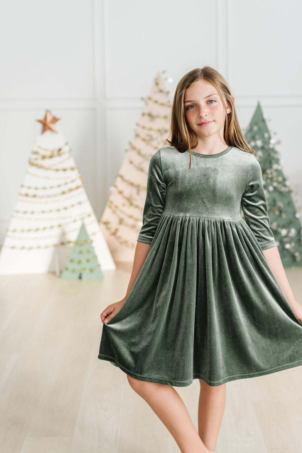 Sage Velvet Twirl Dress by Mila & Rose, featuring a young girl twirling joyfully in a stylish sage green dress, set against a festive backdrop of decorative holiday trees.
