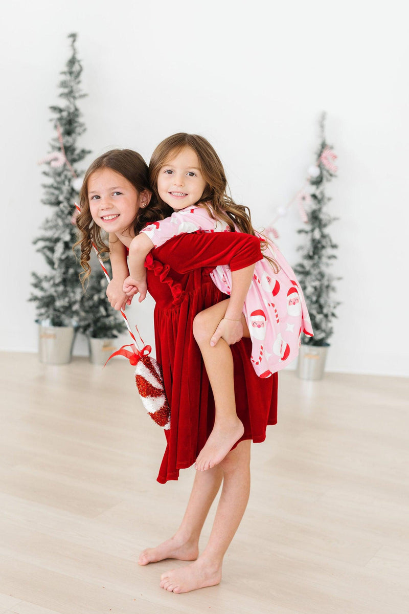 Two girls enjoying the festive season, one in a red Jingle Bells 3/4 Sleeve Pocket Twirl Dress and the other in a pink Santa-patterned dress, perfect for holiday celebrations.