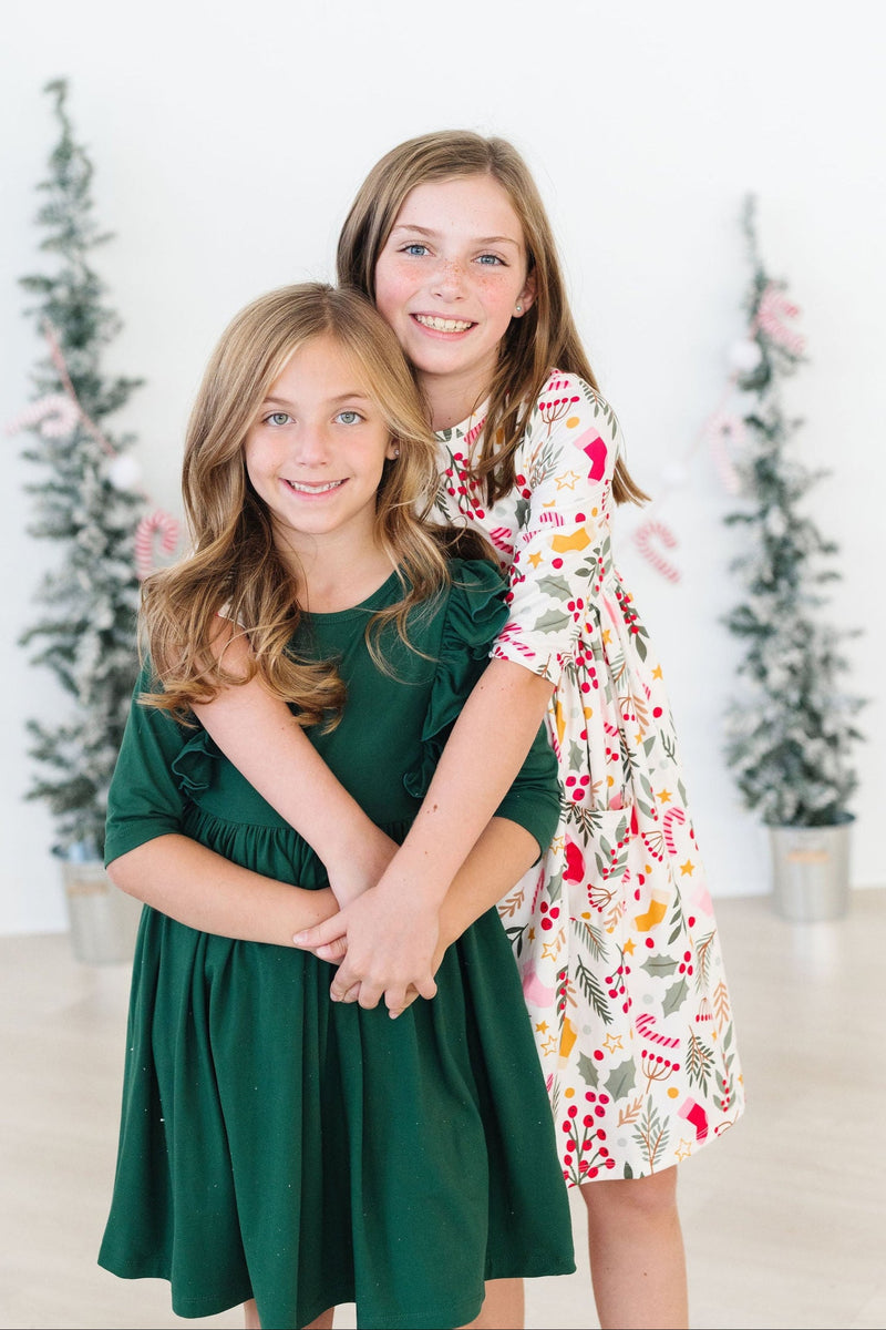 Two young girls hugging, one wearing the 'Holly Jolly Christmas' 3/4 Sleeve Pocket Christmas Twirl Dress for Girls, set against a festive backdrop.