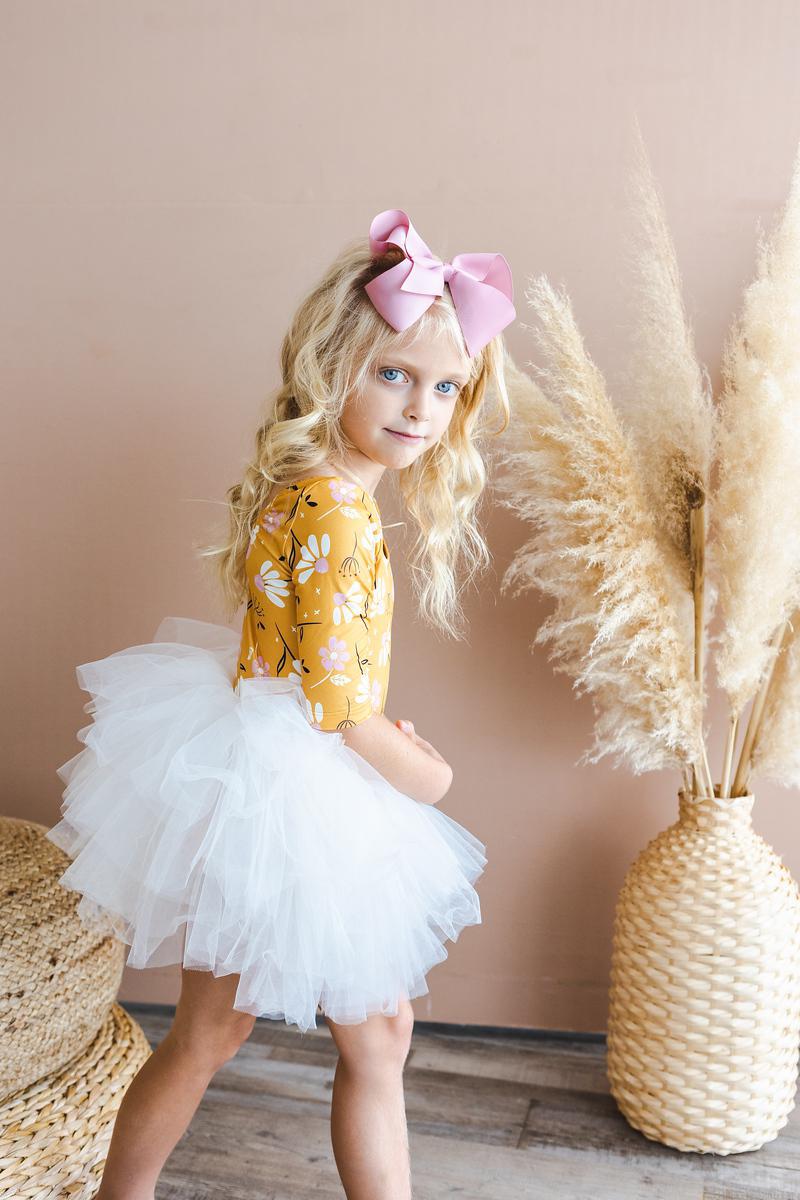 Young girl wearing the Dandelions in Fall 3/4 Tutu Leotard by Mila & Rose® with a fluffy white tutu and a pink bow, posing near decorative plants.