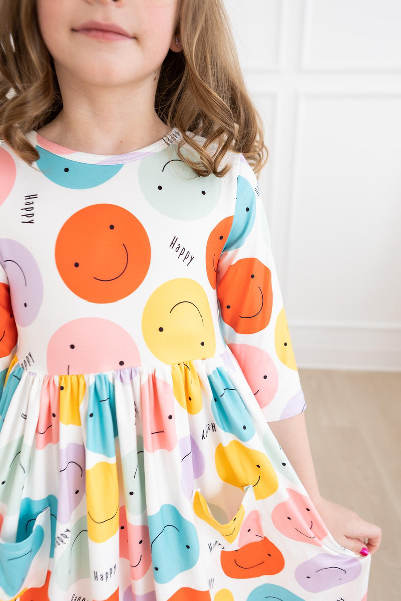 A close-up of a girl in the 'Happy Days' 3/4 Pocket Fun Twirl Dress for Girls, showcasing the playful smiley face patterns and pockets.