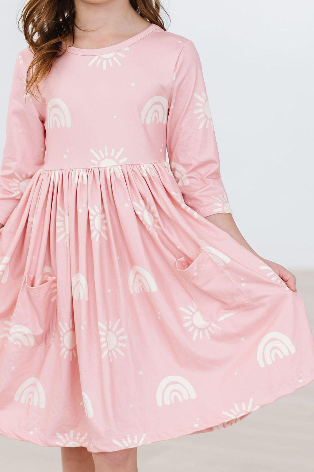 Soft Pink Suns 3/4 Sleeve Pocket Twirl Dress showcasing a girl twirling in a playful pose, featuring rainbow and sun patterns.