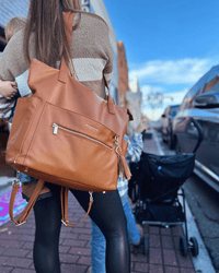 Woman carrying cognac diaper bag on one shoulder in urban setting, showcasing spacious tote silhouette and gold hardware