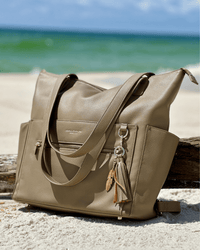 Taupe diaper bag resting on white sand near driftwood with ocean background and gold feet visible