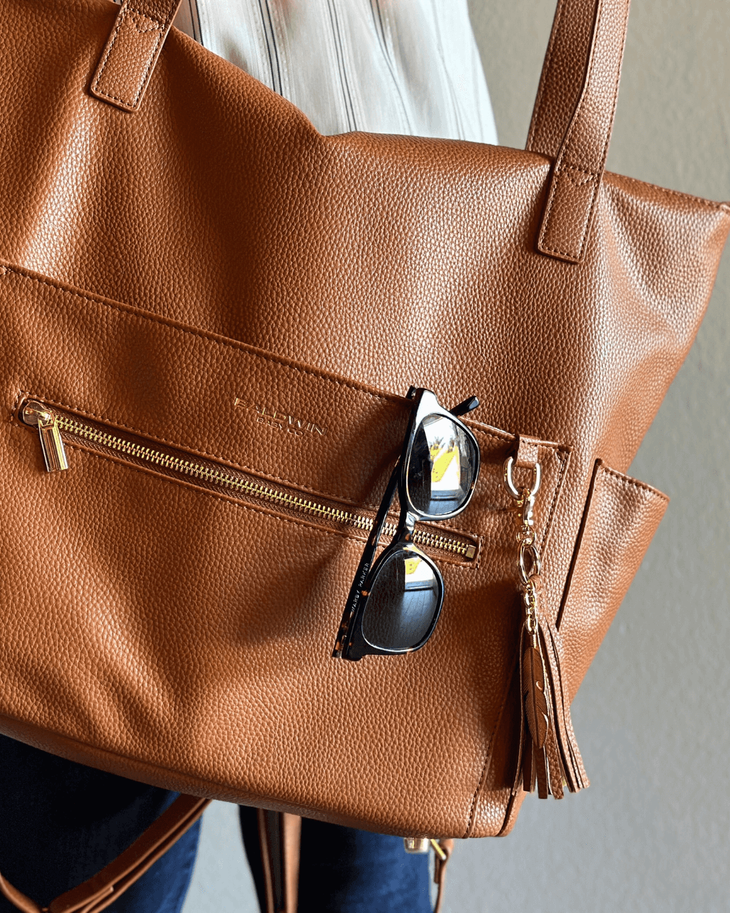 Close-up of cognac pebbled vegan leather diaper bag with gold hardware, feather tassel, and sunglasses tucked in front pocket