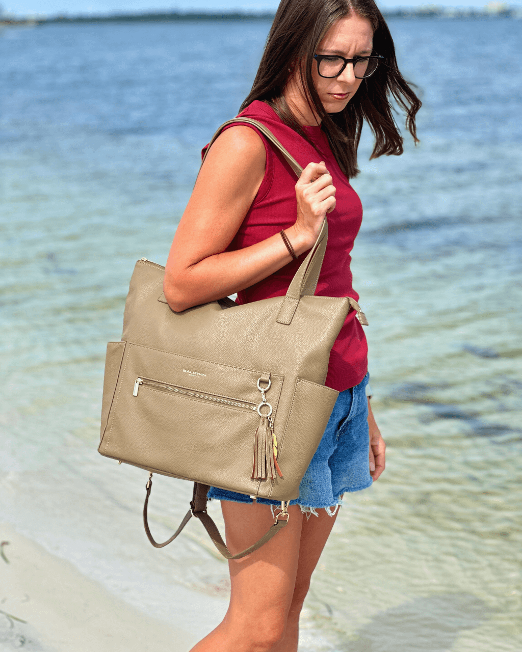 Woman standing by water wearing large taupe diaper bag tote on shoulder with hanging backpack straps