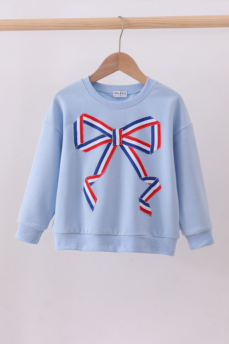 Light blue sweatshirt with a large red, white, and blue bow design on a wooden hanger against a light gray background.