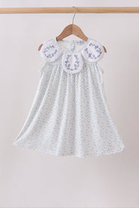 White dress with floral embroidery on a wooden hanger against a beige background