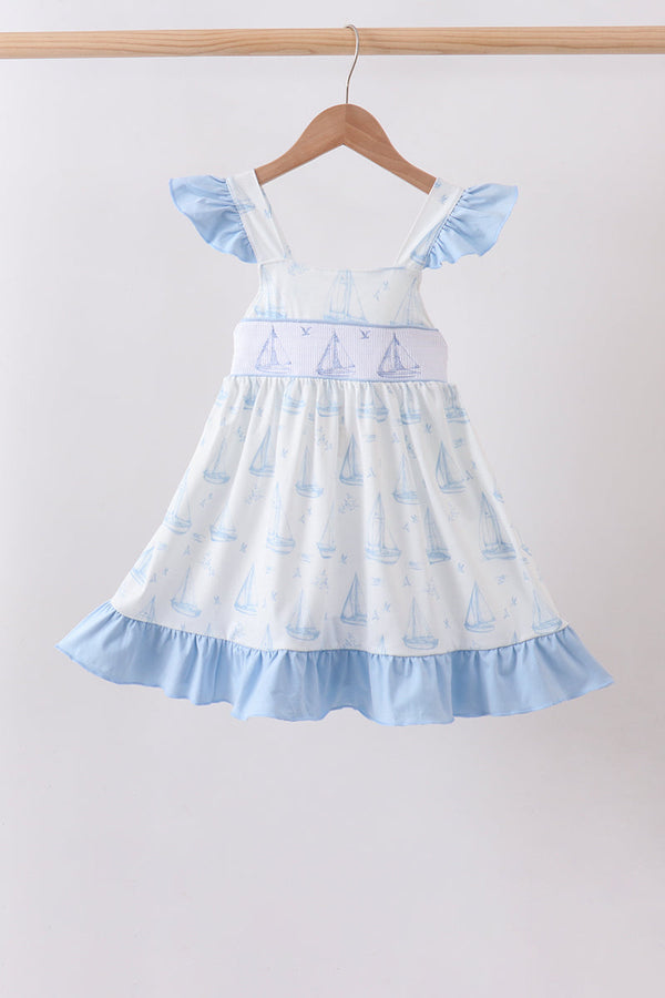 A light blue and white baby girl's dress with a sailboat pattern, featuring ruffled cap sleeves, a smocked bodice, and a ruffled hem, hanging on a wooden hanger against a plain light gray wall.