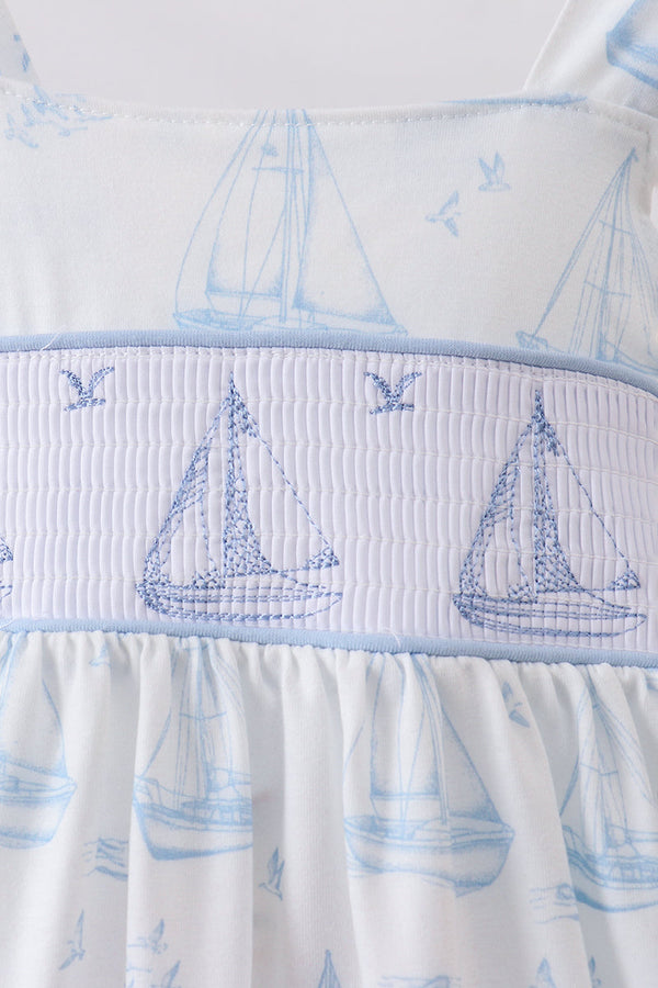 Close-up of a white baby dress with a light blue nautical sailboat pattern and a smocked waistband featuring embroidered sailboats and seagulls.