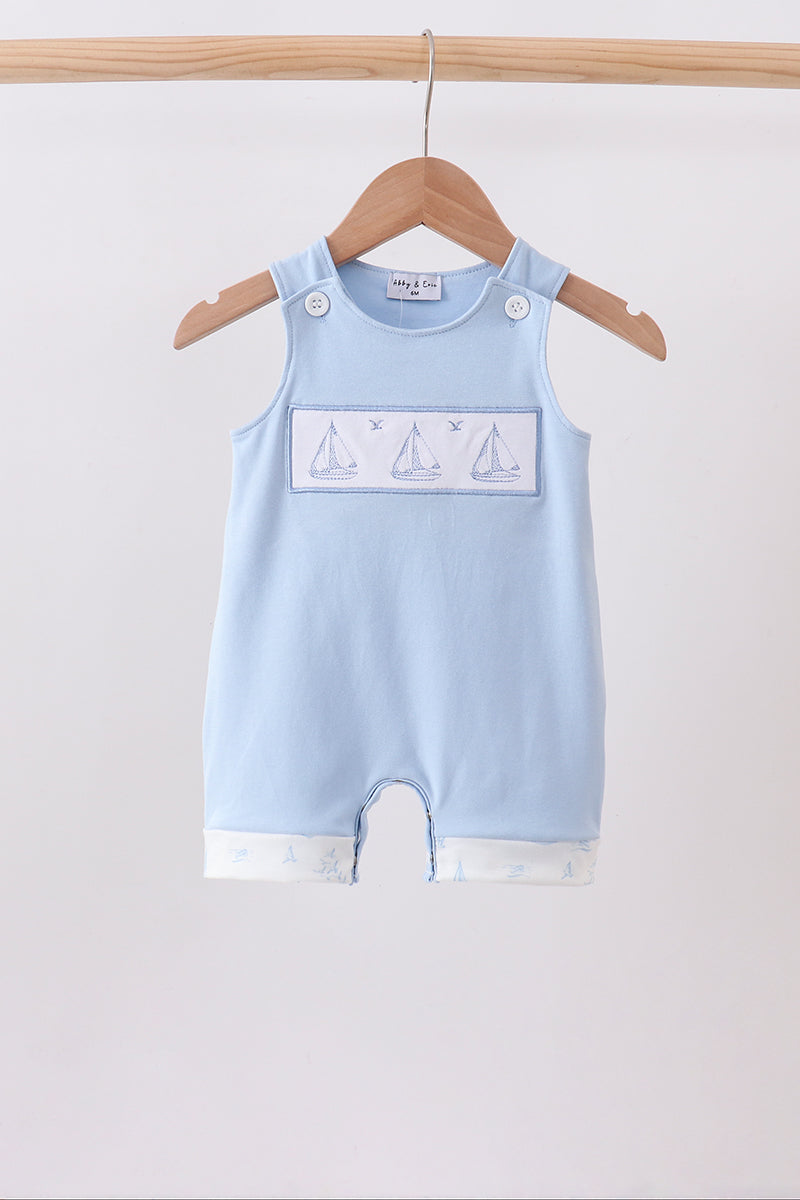 A light blue baby romper with a white rectangular chest panel featuring sailboat illustrations, hanging on a wooden hanger from a wooden clothes rack against a plain white background.