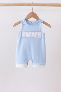 A light blue baby romper with a white rectangular chest panel featuring sailboat illustrations, hanging on a wooden hanger from a wooden clothes rack against a plain white background.