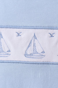 Close-up of a light blue fabric product featuring a white horizontal band with embroidered blue sailboats and birds.