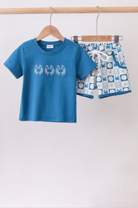 A two-piece kids' outfit set hanging on wooden hangers from a wooden rod. The set includes a short-sleeve crewneck t-shirt in medium blue with three white crab graphics on the chest, and matching patterned shorts in blue, white, and light gray with a white drawstring.