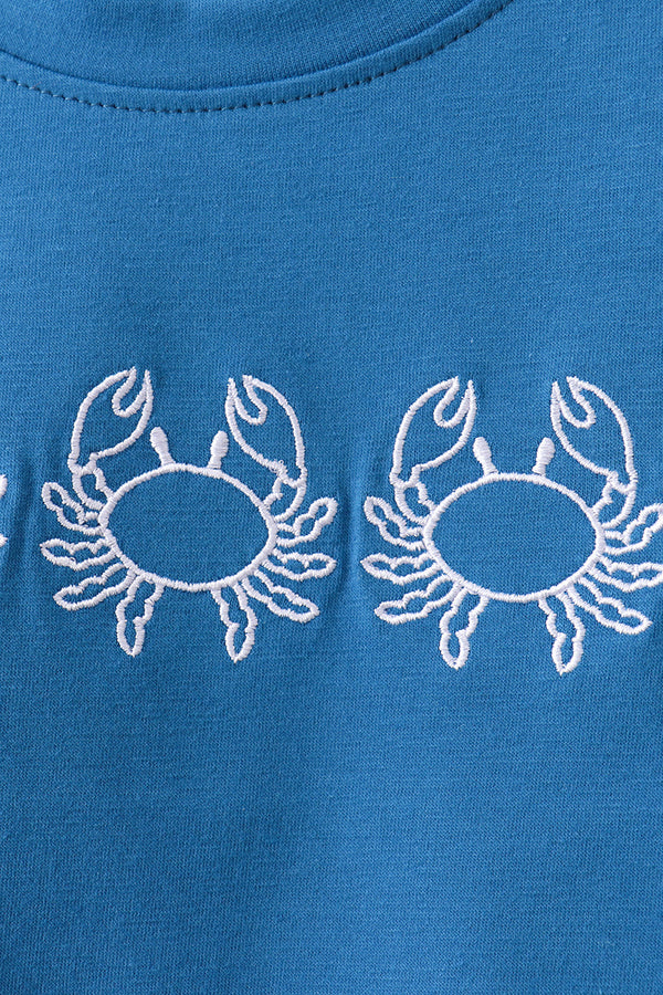 A close-up of a bright blue fabric garment featuring two white embroidered crab motifs across the chest.