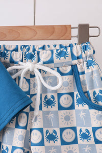 A close-up of a pair of children's shorts hanging on a wooden hanger. The shorts feature a blue and white checkerboard pattern with crabs, smiley faces, palm trees, and suns, with a white drawstring and a solid blue side pocket.
