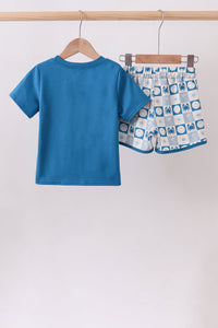 A two-piece children's outfit hanging on wooden hangers from a light wood rod. The set includes a solid bright blue short-sleeve t-shirt and matching patterned shorts with a white base, blue borders, and blue geometric blocks.