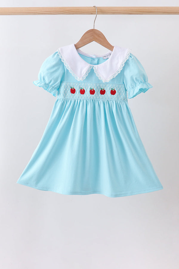 A light blue baby girl's dress with a white Peter Pan collar and embroidered red apple details, hanging on a wooden hanger against a plain white wall.