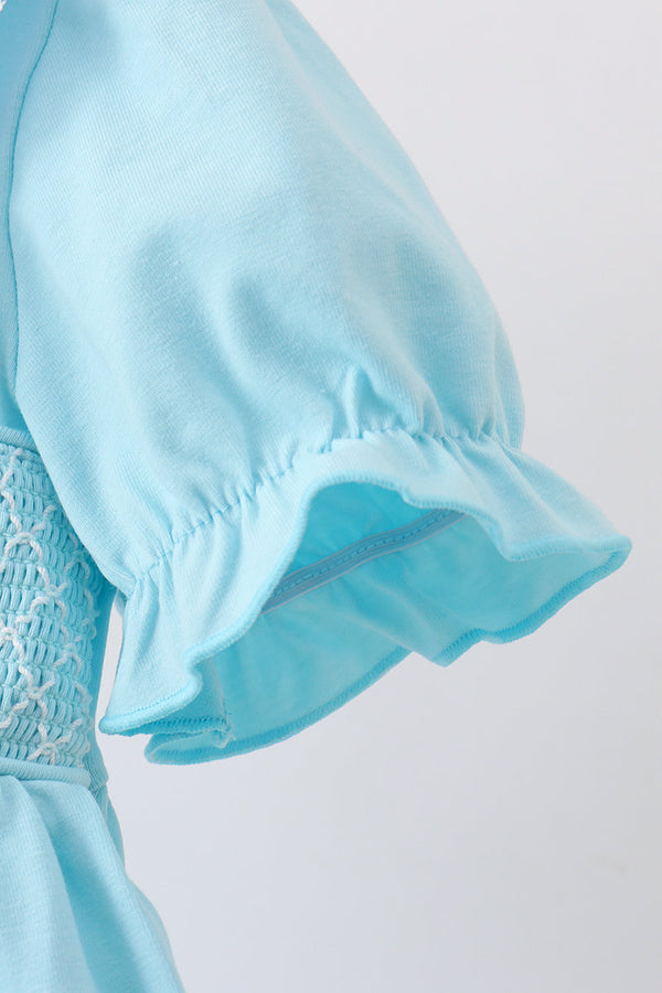 Close-up of a light blue garment sleeve with a ruffled hem and a smocked lace panel at the waist, shown against a plain light background.
