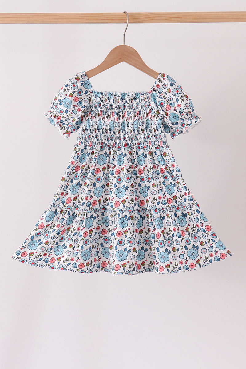 Blue floral smocked ruffle dress for girls with puffed sleeves and delicate bodice detailing on a wooden hanger.