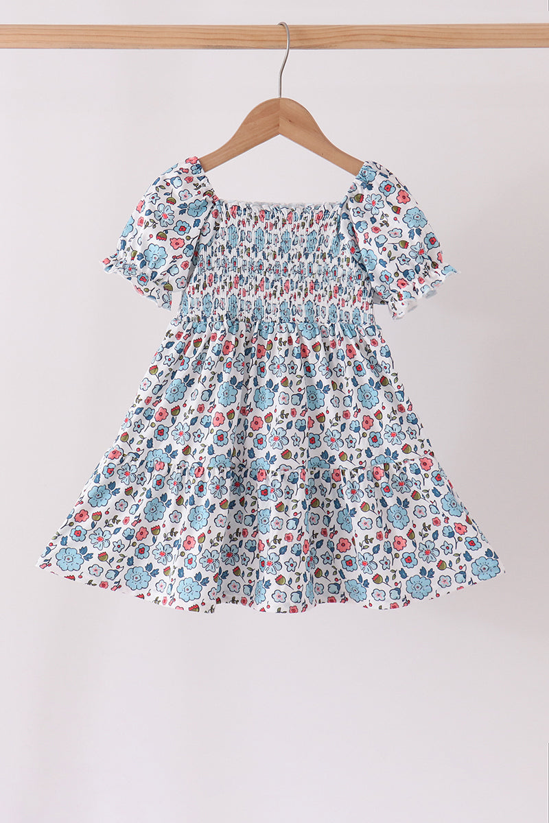 Blue floral girls dress with smocked bodice, ruffled sleeves, and tiered hem hanging on a wooden hanger.