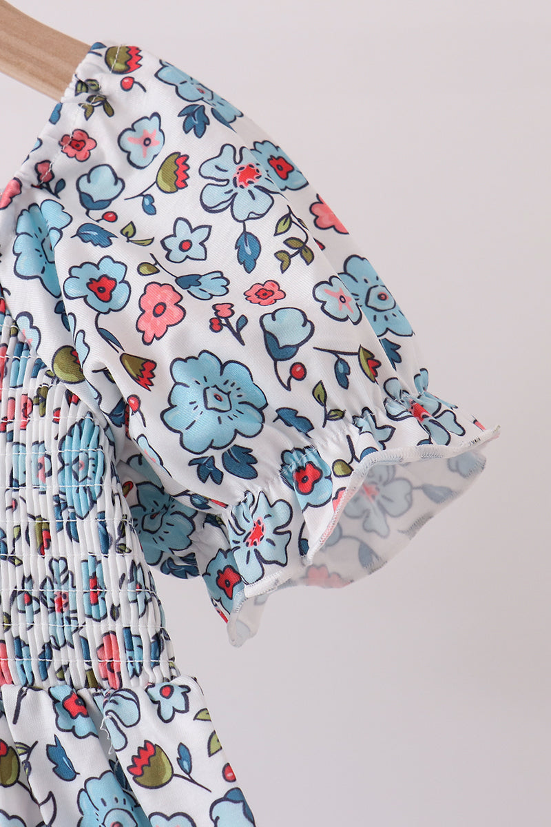 Blue floral girls dress featuring delicate smocked bodice detailing and a ruffled puff sleeve with soft pink and red accents on a white background.