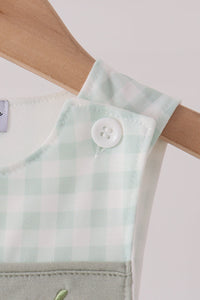 Close-up of sage green gingham romper strap on wooden hanger with white button and pumpkin embroidery detail for fall.