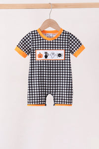 A baby short-sleeve romper with a black and white gingham pattern, orange trim, and a Halloween-themed chest graphic featuring a pumpkin, cat, ghost, and spider, hanging on a wooden hanger.